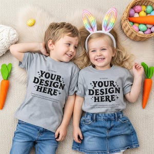 May include: Two children wearing gray t-shirts with the text "YOUR DESIGN HERE" and blue denim bottoms. One child wears a bunny ear headband. Easter basket with colorful eggs and carrot decorations.