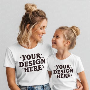 May include: Two white t-shirts with the text "YOUR DESIGN HERE" in dark brown. The shirts are worn by a woman and a young girl, both smiling. The woman and girl are wearing blue jeans. The shirts have a crew neck.