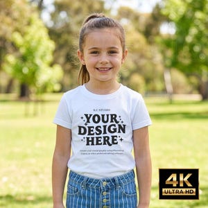 May include: A young person wearing a white t-shirt with the text "YOUR DESIGN HERE" in black. The shirt also has the text "K.C STUDIO" above the main text and a line of text below. The person is wearing blue jeans and is standing outside.