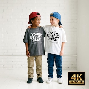 Op de afbeelding: Twee kinderen dragen T-shirts met de tekst "YOUR DESIGN HERE". Eén kind draagt een grijs shirt, een kaki broek en een rode pet. Het andere kind draagt een wit shirt, een blauwe broek en een blauwe pet. Beiden lachen.