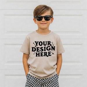Könnte beinhalten: Ein beiges Kurzarm-T-Shirt mit dem schwarzen Text "YOUR DESIGN HERE". Das Kind trägt eine schwarze Sonnenbrille und karierte Shorts. Das Shirt besteht aus einem weichen Material.