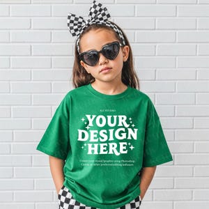 May include: A child wearing a green t-shirt with the text "YOUR DESIGN HERE" in white. They are also wearing heart-shaped sunglasses, a black and white checkered headband, and checkered trousers.