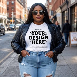 May include: A woman wearing a black leather jacket, sunglasses, and light blue jeans. She is wearing a white t-shirt with the text "YOUR DESIGN HERE" in a bold font. The background shows a city street.