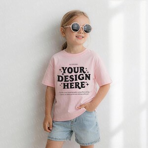 Op de afbeelding: Een jong persoon draagt een lichtroze t-shirt en lichtblauwe spijkerbroekshorts. Op het t-shirt staat de tekst "YOUR DESIGN HERE" in het zwart. De persoon draagt ook een ronde, lichtgekleurde zonnebril.