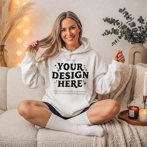 Pode incluir: Um moletom branco com o texto "YOUR DESIGN HERE" em negrito preto. O moletom tem um bolso frontal e um capuz com cordão. A imagem também inclui uma mulher sentada em um sofá.