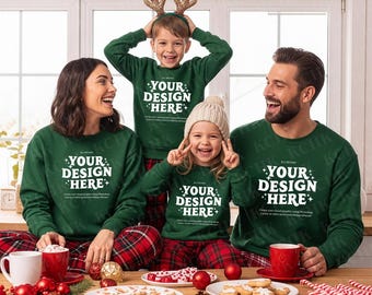 May include: A family of four wearing matching green sweatshirts and red plaid pajamas. The sweatshirts have the text "YOUR DESIGN HERE+". The family is smiling and gathered around a table with cookies and Christmas decorations.