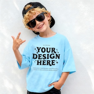 May include: A light blue t-shirt with the text "YOUR DESIGN HERE" in black. The shirt is worn by a person wearing sunglasses and a black cap. The text "Create your visual/graphic using Photoshop, Canva, or other preferred editing software" is also visible.