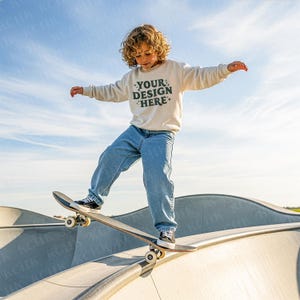 May include: A child wearing a cream-colored sweatshirt and blue jeans balances on a skateboard at a skate park. The sweatshirt has the text "YOUR DESIGN HERE" in dark green. The skateboard is black and gray.