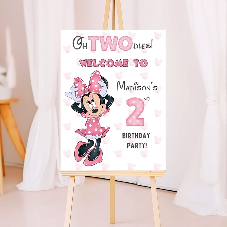May include: A white sign on an easel with the text "Oh TWO-dles! Welcome to Madison's 2nd Birthday Party!" A cartoon Minnie Mouse in a pink polka dot dress and bow is featured on the sign. Pink heart shapes are scattered around the sign.