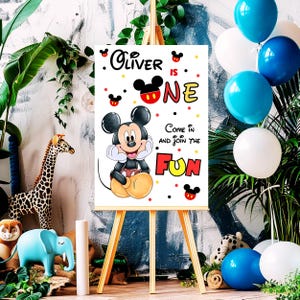 May include: A white sign on a wooden easel featuring Mickey Mouse and the text "Oliver is One." The sign includes the phrase "Come in and join the fun" and is decorated with red, yellow, and black accents. Balloons in blue and white are in the background.