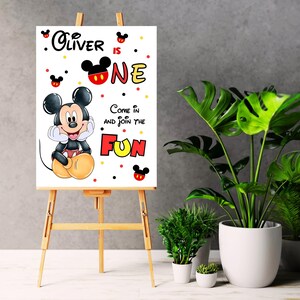 May include: A white sign on a wooden easel featuring Mickey Mouse and the text "Oliver is One." The sign has a cartoon illustration of Mickey Mouse and the words "Come in and join the FUN" in red and yellow.