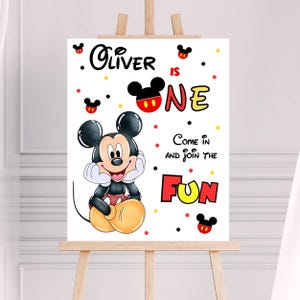 May include: A white sign on an easel featuring Mickey Mouse. The sign reads "Oliver is One" with the words "Come in and Join the Fun." Mickey Mouse is in the center, surrounded by red, yellow, and black accents.