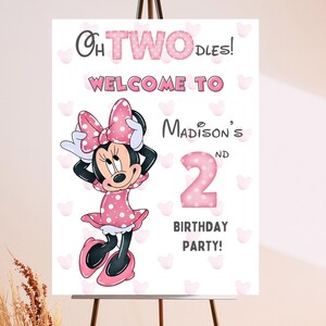 May include: A birthday party sign featuring Minnie Mouse. The sign reads "Oh TWO-dles! Welcome to Madison's 2nd Birthday Party!" Minnie Mouse is wearing a pink polka dot dress and bow. The background is white with pink heart shapes.
