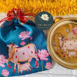 May include: A teal drawstring bag and a gold-rimmed plate with cow illustrations. The bag features a cow and lotus flower design. The plate has a cow and calf illustration. The bag has a red drawstring.
