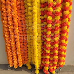 Puede incluir: Una colección de guirnaldas de caléndulas en colores vibrantes. Las guirnaldas son en tonos naranja, amarillo y una mezcla de rojo y amarillo. Cada guirnalda está hecha de flores de caléndula redondas y densamente empaquetadas, creando una apariencia completa y texturizada. Las guirnaldas cuelgan verticalmente.