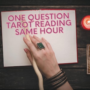 May include: A white notebook with the text "ONE QUESTION TAROT READING SAME HOUR" written in pink. A hand with a green gemstone ring is holding a wooden stick. The notebook is surrounded by candles and other objects.