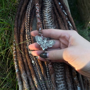 May include: A silver butterfly charm with intricate details hangs from a set of brown and gray dreadlocks. The dreadlocks are decorated with small silver beads and a brown and white braided cord.