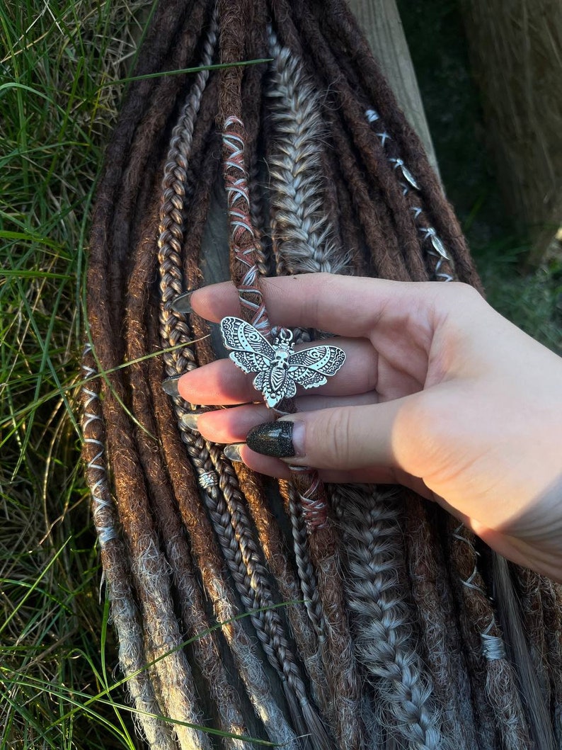 May include: A silver butterfly charm with intricate details hangs from a set of brown and gray dreadlocks. The dreadlocks are decorated with small silver beads and a brown and white braided cord.