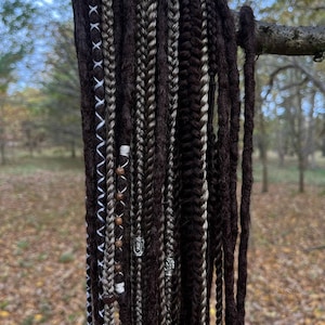 May include: A collection of brown and grey synthetic dreadlocks with white and brown beads and silver rings hanging from a branch.