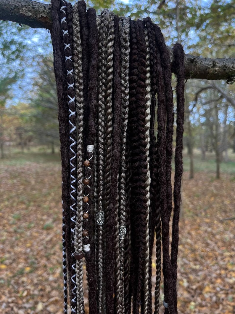 May include: A collection of brown and grey synthetic dreadlocks with white and brown beads and silver rings hanging from a branch.