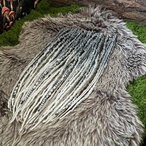 May include: A collection of gray and white dreadlocks with decorative patterns, arranged on a faux fur surface. The dreadlocks are a mix of solid and patterned strands, creating a textured appearance. The background includes green moss and a dark, mushroom-themed fabric.
