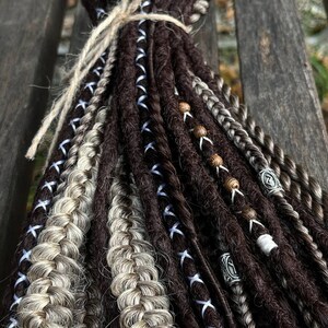 May include: A bundle of brown and blonde synthetic dreadlocks with blue and white ribbon accents. The dreadlocks are tied together with twine and have wooden and metal beads.