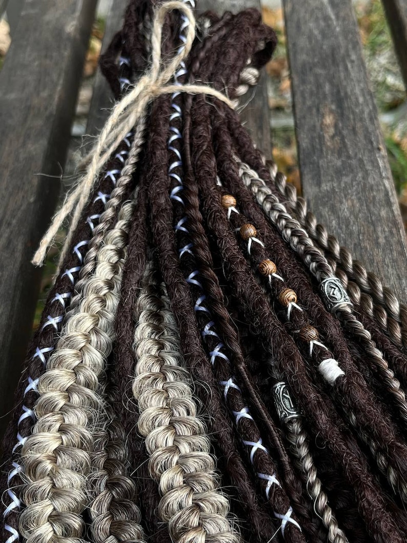 May include: A bundle of brown and blonde synthetic dreadlocks with blue and white ribbon accents. The dreadlocks are tied together with twine and have wooden and metal beads.