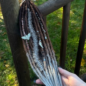 May include: Brown and white synthetic dreadlocks with silver beads and charms, including a butterfly charm. The dreadlocks are tied together with a brown string.