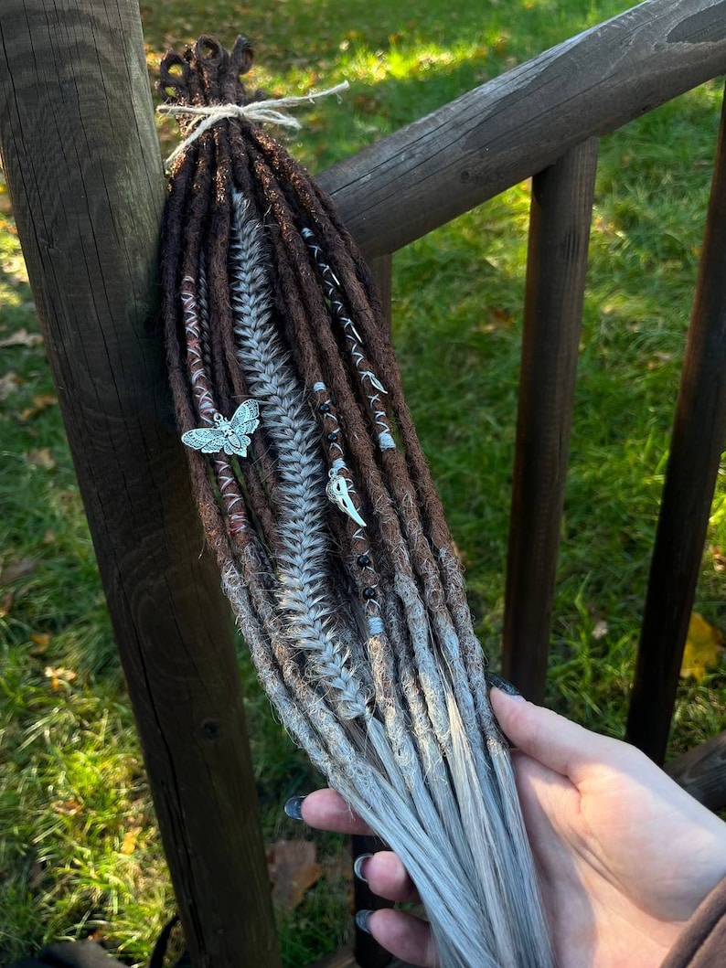 May include: Brown and white synthetic dreadlocks with silver beads and charms, including a butterfly charm. The dreadlocks are tied together with a brown string.