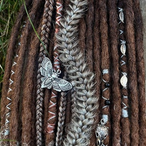 May include: Close-up of brown and grey synthetic dreadlocks with silver butterfly and skull charms. The dreadlocks are decorated with brown and white string and black beads.