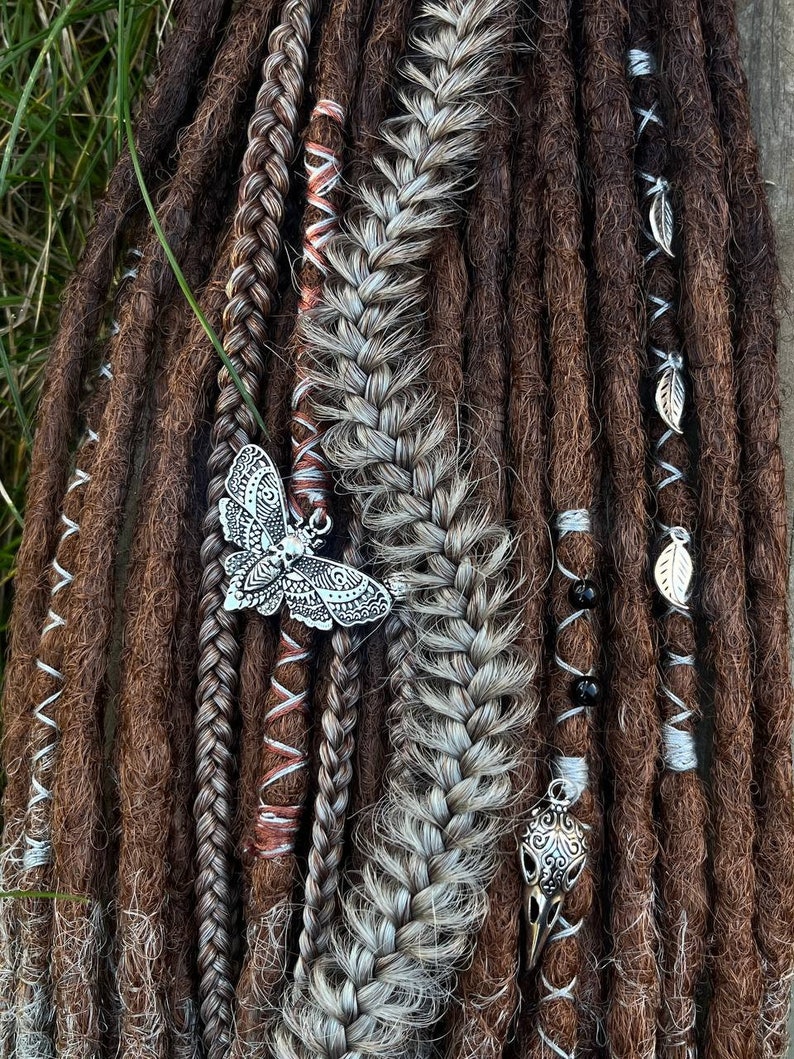 May include: Close-up of brown and grey synthetic dreadlocks with silver butterfly and skull charms. The dreadlocks are decorated with brown and white string and black beads.