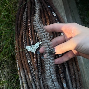 May include: A set of brown and gray synthetic dreadlocks with a silver butterfly charm. The dreadlocks are arranged in a braid with a few strands of brown and gray hair.
