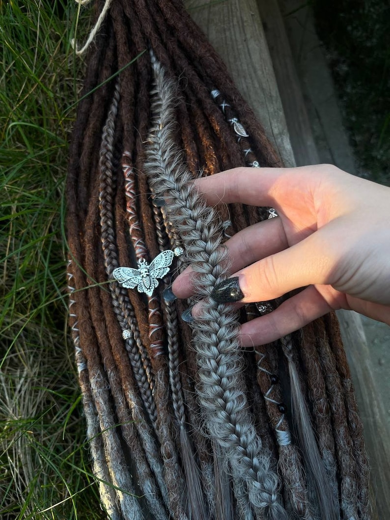 May include: A set of brown and gray synthetic dreadlocks with a silver butterfly charm. The dreadlocks are arranged in a braid with a few strands of brown and gray hair.