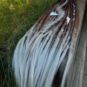 May include: A set of synthetic dreadlocks with brown and gray ombre hair. The dreadlocks are decorated with feathers and beads.