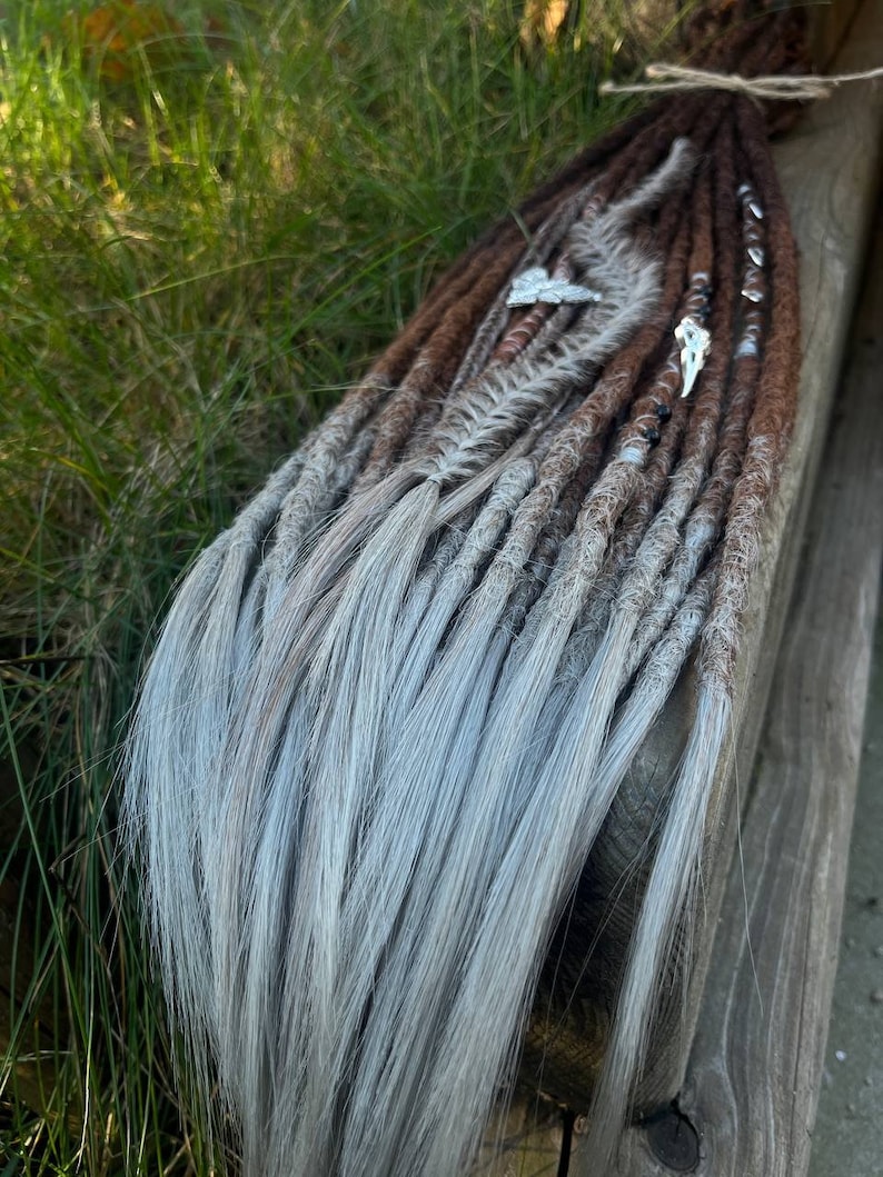 May include: A set of synthetic dreadlocks with brown and gray ombre hair. The dreadlocks are decorated with feathers and beads.