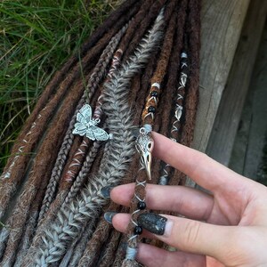 May include: Brown and gray synthetic dreadlocks with silver and black beads and charms. The charms are a butterfly and a bird skull.