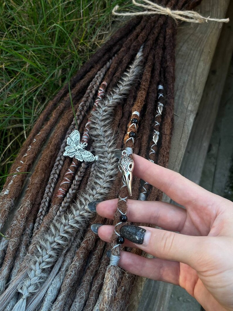 May include: Brown and gray synthetic dreadlocks with silver and black beads and charms. The charms are a butterfly and a bird skull.