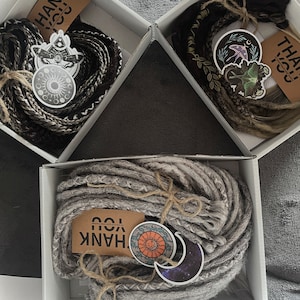 May include: Three white boxes with lids open, each containing a bundle of synthetic hair extensions. The hair is in different colors: black and grey, brown and blonde, and grey. Each box has a thank you tag and a sticker with a celestial or nature-themed design.