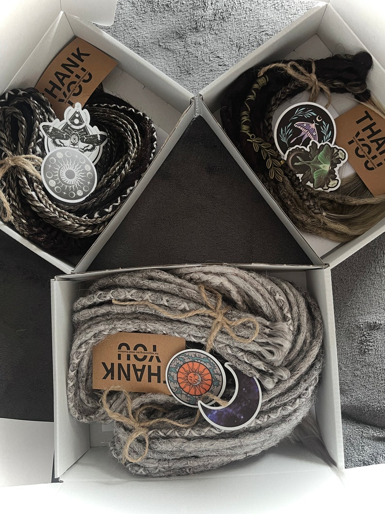 May include: Three white boxes with lids open, each containing a bundle of synthetic hair extensions. The hair is in different colors: black and grey, brown and blonde, and grey. Each box has a thank you tag and a sticker with a celestial or nature-themed design.