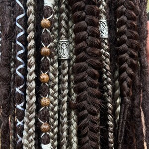 May include: Close-up of a variety of dreadlocks with different textures and colours, including black, grey, and white. Some dreadlocks have wooden beads and silver metal beads with rune symbols.