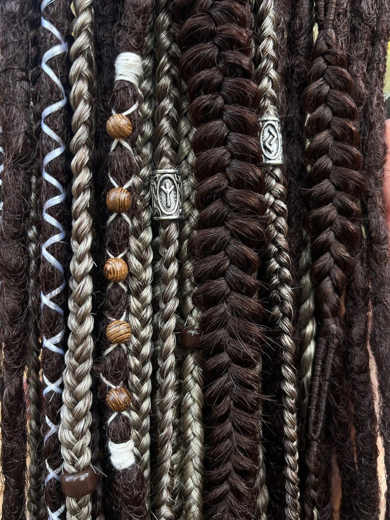 May include: Close-up of a variety of dreadlocks with different textures and colours, including black, grey, and white. Some dreadlocks have wooden beads and silver metal beads with rune symbols.