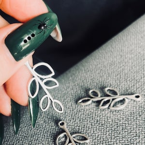 May include: Three silver leaf charms with a hole at the top for attaching to jewelry or other crafts. The charms are in the shape of a branch with five leaves each.