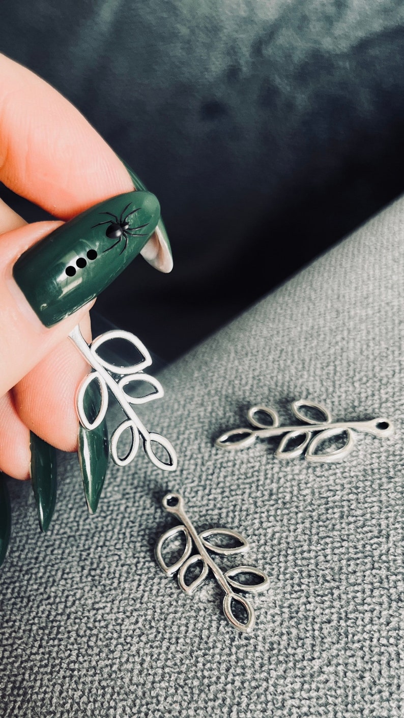 May include: Three silver leaf charms with a hole at the top for attaching to jewelry or other crafts. The charms are in the shape of a branch with five leaves each.