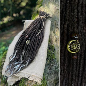 May include: A bundle of dark brown dreadlocks with white-tipped ends, tied with twine. The dreadlocks are decorated with wooden beads and a gold-toned metal charm featuring a sun and moon face design, visible in a detailed close-up view.
