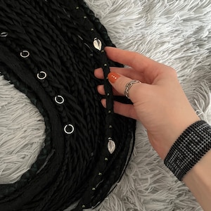 May include: Black synthetic hair extensions with small silver rings and leaf-shaped charms. The hair is braided and has a textured look.