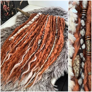 Op de afbeelding: Een set oranje en witte dreadlocks met decoratieve kralen en leren accenten. De dreadlocks zijn gerangschikt op een imitatiebont oppervlak, dat de textuur en kleurvariaties laat zien. Sommige dreadlocks zijn versierd met kralen en leren wikkelingen.