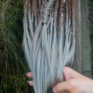 May include: A set of brown and grey synthetic dreadlocks with silver beads and charms. The dreadlocks are ombre, transitioning from brown to grey.