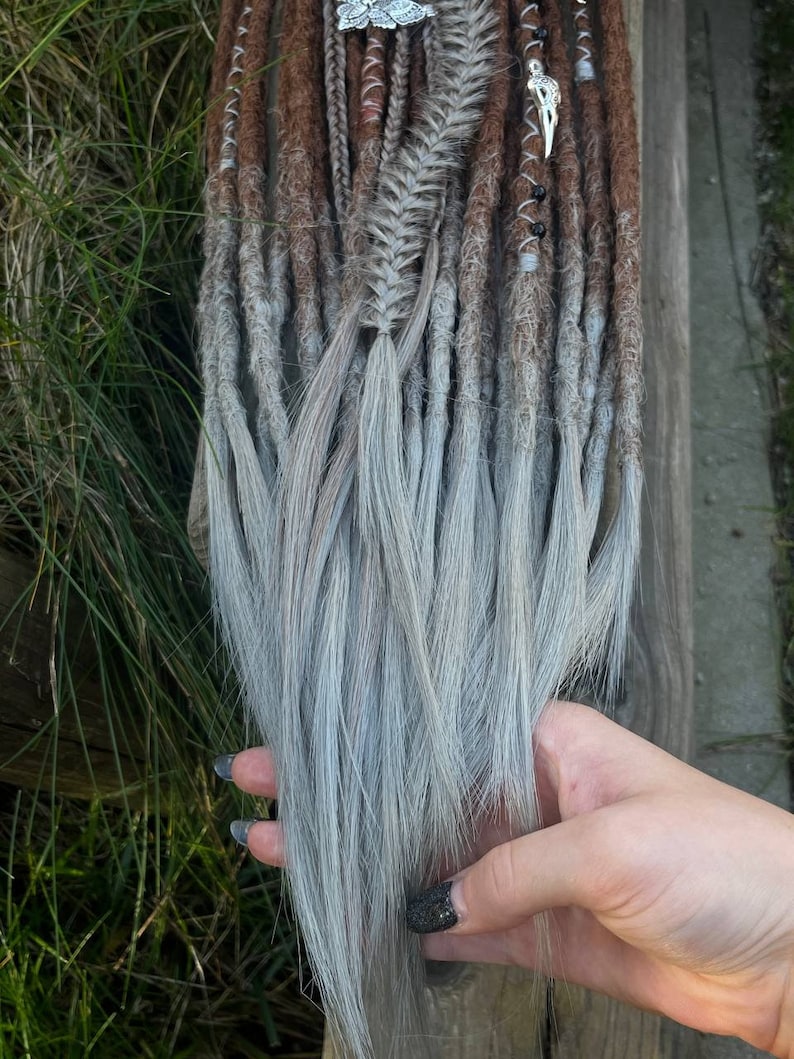 May include: A set of brown and grey synthetic dreadlocks with silver beads and charms. The dreadlocks are ombre, transitioning from brown to grey.
