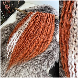 May include: A bundle of orange and white curly hair extensions. The extensions are arranged on a gray faux fur surface. The hair is a vibrant orange with a white stripe running through the middle. The hair is styled in a wavy, curly pattern.