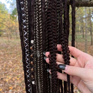 May include: A collection of brown, black and grey synthetic dreadlocks with beads and metal charms. The dreadlocks are hanging from a branch.